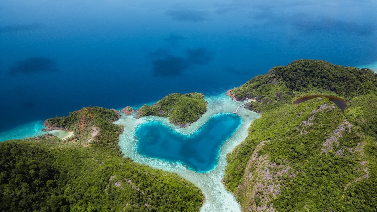 Heart shaped Love Lagoon seen from a cliff viewpoint in Misool, Raja Ampat, with turquoise water surrounded by limestone formations.